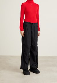 Persona che indossa un maglione a collo alto rosso, pantaloni neri larghi con tasche laterali e sneakers platform nere, in piedi su un pavimento beige.
