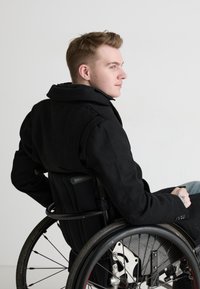 Black coat with a high collar, seated in a wheelchair, featuring a textured fabric and prominent stitching details. Wheels with red accents.