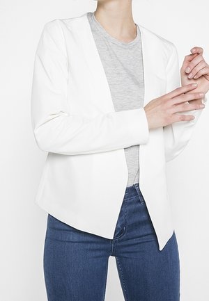 Femme portant un blazer blanc ouvert sur un t-shirt gris et un jean bleu, les mains se touchant doucement.