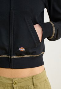 Μαύρο φούτερ με φερμουάρ από μείγμα βαμβακιού, με ραβδωτές μανσέτες, χρυσές ραφές και ορατή ετικέτα μπροστά. Τσέπες και στις δύο πλευρές.