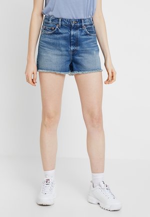 Person iført midtaljede blå denimshorts med frynser i kanterne, hvide sokker og hvide sneakers, stående foran en ensartet baggrund.
