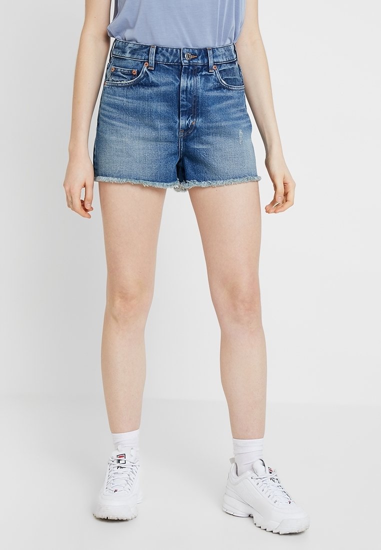 Personne portant un short en denim bleu taille moyenne avec des ourlets effilochés, des chaussettes blanches et des baskets blanches, se tenant devant un fond uni.