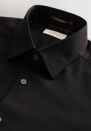 Chemise noire à col, fabriquée en tissu texturé. Présente un design classique avec deux boutons gris et une étiquette dans le col, ornée d'accents marron.