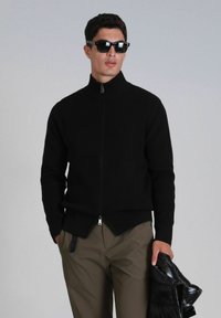 Pull zippé noir côtelé avec un col haut, doté d'un ourlet fendu, associé à un pantalon marron clair et une doudoune noire brillante.