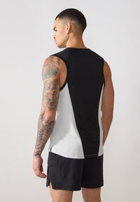 Schwarz-weißes ärmelloses Sportoberteil mit einem strukturierten schwarzen Rückenteil und geschwungenen weißen Akzenten, kombiniert mit schwarzen Shorts, die Armtätowierungen zeigen.