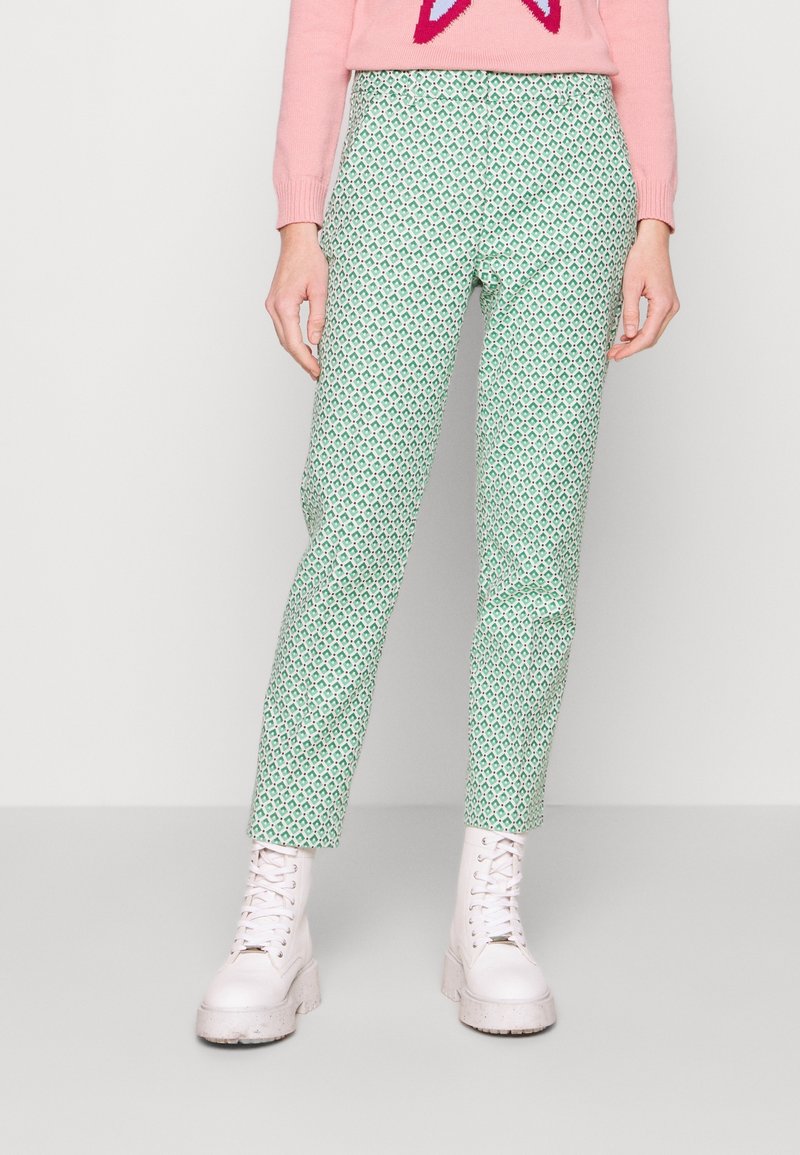 Pantalons verts à motif avec une coupe slim, mettant en avant un design géométrique. Portés avec un pull rose et des bottes blanches épaisses.
