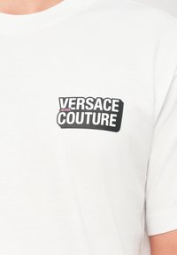 T-shirt branca com o logótipo "Versace Jeans Couture" em preto e rosa impresso no peito. Apenas a parte superior do torso visível.