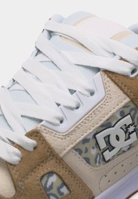 Αθλητικό παπούτσι με λευκή δαντέλα, επενδύσεις από μπεζ και καφέ δέρμα σουέντ, λεπτομέρεια από ύφασμα καμουφλάζ και λογότυπο DC στο πλάι.