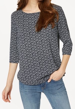 Mujer con una blusa de manga tres cuartos con estampado en blanco y negro, pantalones vaqueros azules, una mano en el bolsillo y el cabello castaño suelto sobre el hombro.
