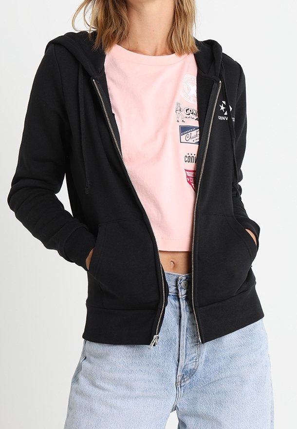Personne portant un sweat à capuche noir zippé par-dessus un t-shirt court rose à motif graphique et un jean taille haute bleu clair, mains dans les poches.