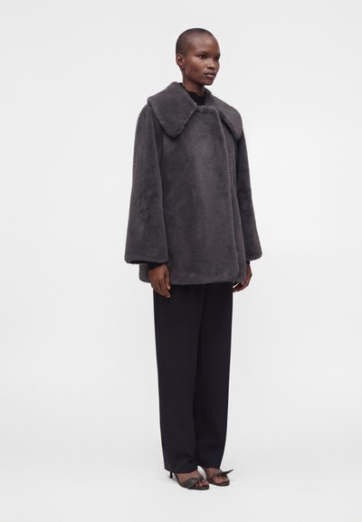 Personne portant un manteau en fausse fourrure gris foncé oversize, un pantalon noir à jambes larges et des talons ouverts noirs, debout devant un fond blanc uni.