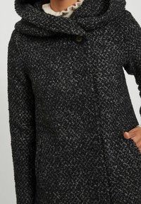 Svart och grå texturerad kappa med stor huva, framknäppning med knappar och sidofickor, med ett mönstrat tyg och mjukt foder.