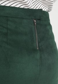 Mörkgrön, mockaliknande kjol med dragkedja på sidan, som har en slät yta och rena sömmar nära sömmen.