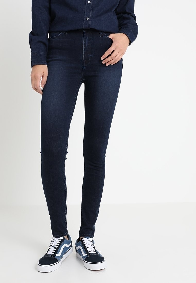 Jeans skinny azul oscuro de mezclilla, con una cintura alta y una textura suave, combinados con zapatillas navy y blancas con un diseño a rayas.