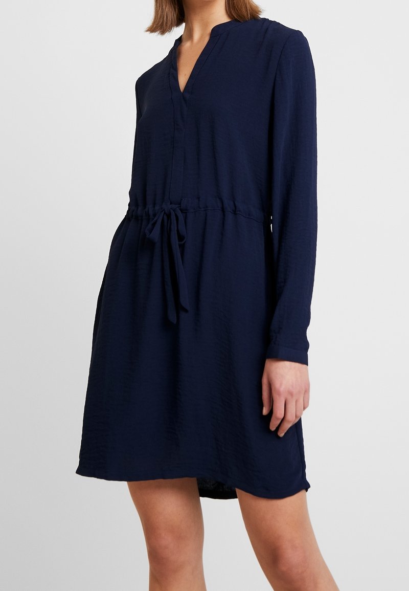 Robe longue bleu marine à manches longues avec un décolleté en V, une taille ajustable par cordon et un tissu texturé. Elle présente une coupe décontractée et un ourlet légèrement évasé.