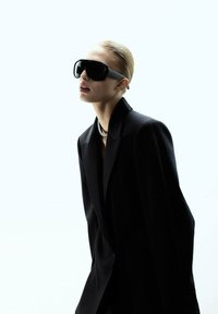 Crne velike sunčane naočale s mat sivoj traci, u paru sa strukturiranim crnim sakoom. Pozadina je svijetla i minimalna.