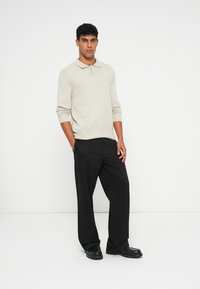 Polo bege de mangas longas com colarinho abotoado, combinado com calças pretas largas e calçado preto. Textura suave e design minimalista.