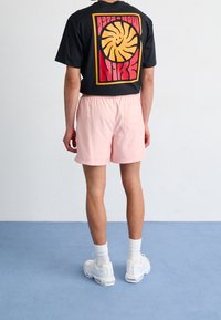 Person som bär en svart T-shirt med en färgglad solgrafik och "Nike"-text, rosa shorts, vita strumpor och vita sneakers som står på ett blått golv.