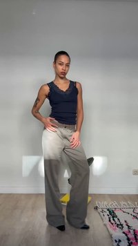 Νεαρό άτομο με τατουάζ ποζάρει σε εσωτερικό χώρο φορώντας μπλε δαντελένιο τοπ με τιράντες και φαρδιά μπεζ παντελόνια, όρθιο σε ξύλινο πάτωμα κοντά σε ένα μοτίβο χαλί.