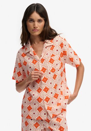 Femme portant une chemise à manches courtes boutonnée et un short assorti avec un motif géométrique orange et rouge, posant avec une main dans la poche.