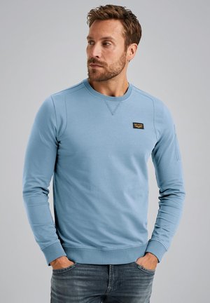 Svetlo modra trenerka z dolgimi rokavi, krožnim ovratnikom, rebrastimi manšetami in robom; ima črno oznako na prsih. Gladek material iz mešanice bombaža.