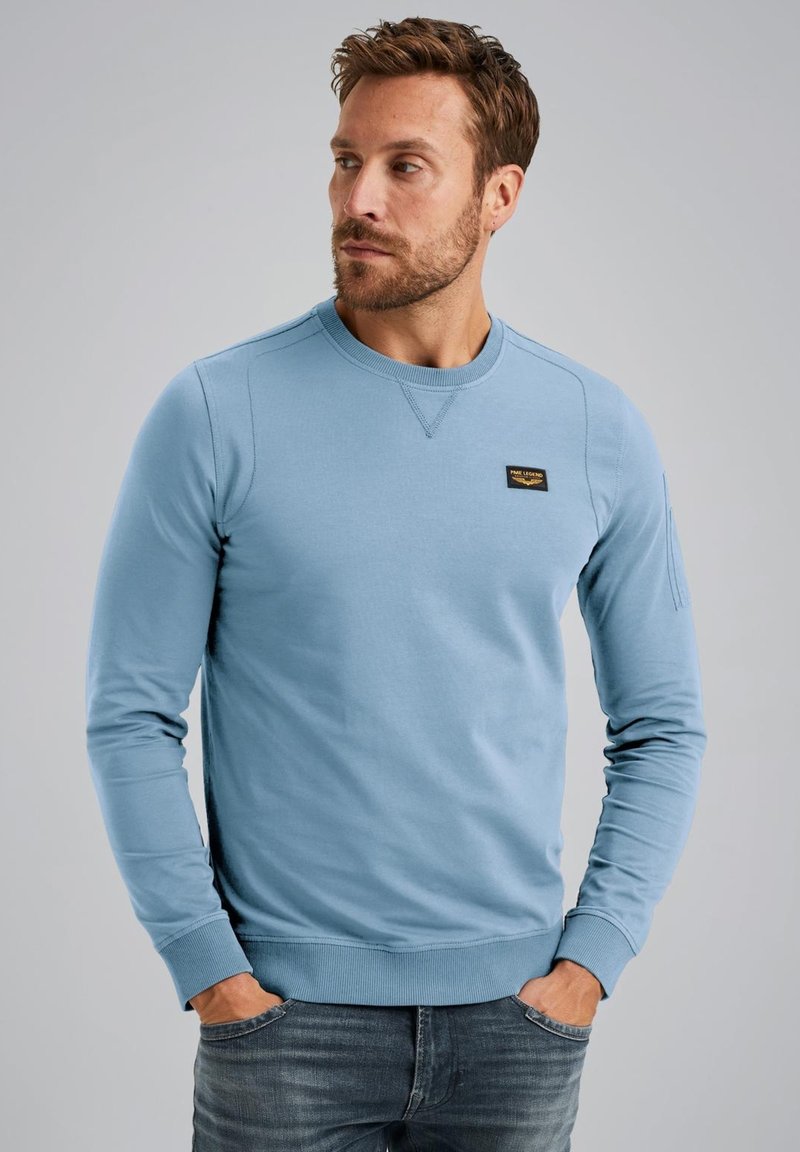 Sweat-shirt bleu clair à manches longues, col rond, avec des poignets et un ourlet côtelés ; présente une étiquette noire sur la poitrine. Tissu en mélange de coton doux.