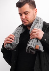 Gray knitted scarf with ribbed texture, showing a leather tag. Held by a person, draped loosely around the neck.