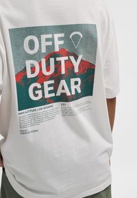 Vit t-shirt med en grafisk tryck av ett rött berg, med texten "OFF DUTY GEAR" och informativ text nedanför i grönt och svart.