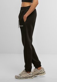 Pantalons de survêtement en coton noir avec une taille élastique, des poches latérales, et un texte brodé "progresize" sur la hanche gauche. Jambes fuselées aux chevilles.