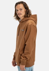 Brown pullover hoodie with a front pocket, drawstring hood, and ribbed cuffs. Features a small logo on the chest. Made of soft fabric.