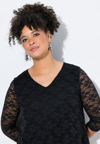 Top in pizzo nero con scollatura a V, maniche lunghe e trasparenti, motivo floreale e vestibilità morbida. Il tessuto presenta un design testurizzato e intricato.