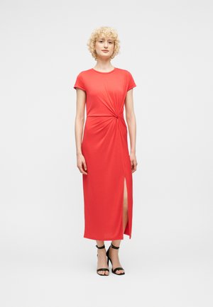 Femme aux cheveux blonds bouclés courts, portant une robe midi corail avec une fente latérale et des sandales noires à talons, debout devant un fond uni.