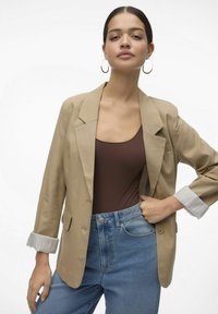 Beige blazer med en skräddarsydd design, med ett slagkrage och randiga manschetter. Bärs över en brun bodysuit och kombineras med högmidjade jeans.