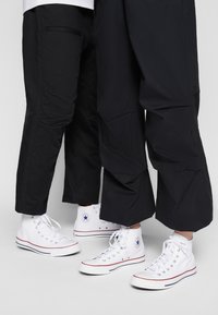 Schwarze tapered Pants mit strukturierter Oberfläche, kombiniert mit weißen High-Top-Sneakern, die ein blaues Stern-Logo und rote Akzente entlang der Sohle aufweisen.