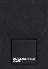 Materiale nero testurizzato con una patch quadrata del logo che presenta la scritta bianca: "KARL LAGERFELD JEANS" su uno sfondo nero liscio.
