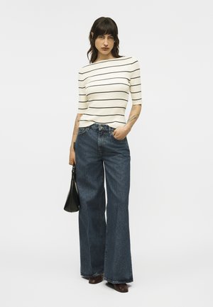 Woman standing with hand in pocket, wearing white short-sleeve top with black stripes, wide-leg dark jeans, brown shoes, and holding a black handbag.