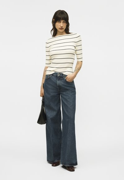 Femme debout, main dans la poche, portant un haut blanc à manches courtes avec des rayures noires, un jean large de couleur foncée, des chaussures marron, et tenant un sac à main noir.