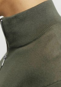 Alyvuogių žalios spalvos užtrauktukas su storu raštu. Artimas vaizdas, parodantis glotnų audinį ir sidabrinį užtrauktuką prie kaklo.