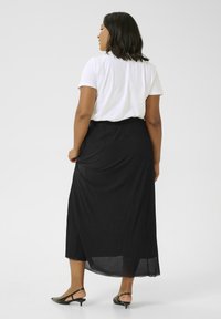 Jupe maxi plissée noire au design fluide, associée à un t-shirt blanc. Portée avec des sandales à talons vertes, affichant un look décontracté.