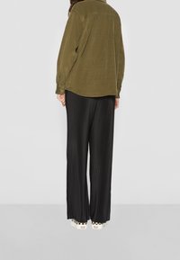 Camicia in corduroy verde oliva con vestibilità rilassata, abbinata a pantaloni a gamba larga neri con pieghe e scarpe slip-on a quadri.