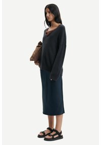 Maglione oversized grigio scuro a maglia con scollatura a V, abbinato a una gonna midi blu navy e sandali neri con cinturini. Tiene una borsa marrone.