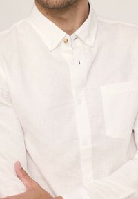 Chemise blanche en lin à col pointu court, avec une petite poche avant et une fermeture à boutons classique avec un bouton contrastant.