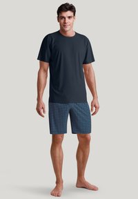 Donkere T-shirt met korte mouwen, gecombineerd met blauwe zwembroek met patronen, featuring geometrische ontwerpen. Model staat blootsvoets tegen een neutrale achtergrond.