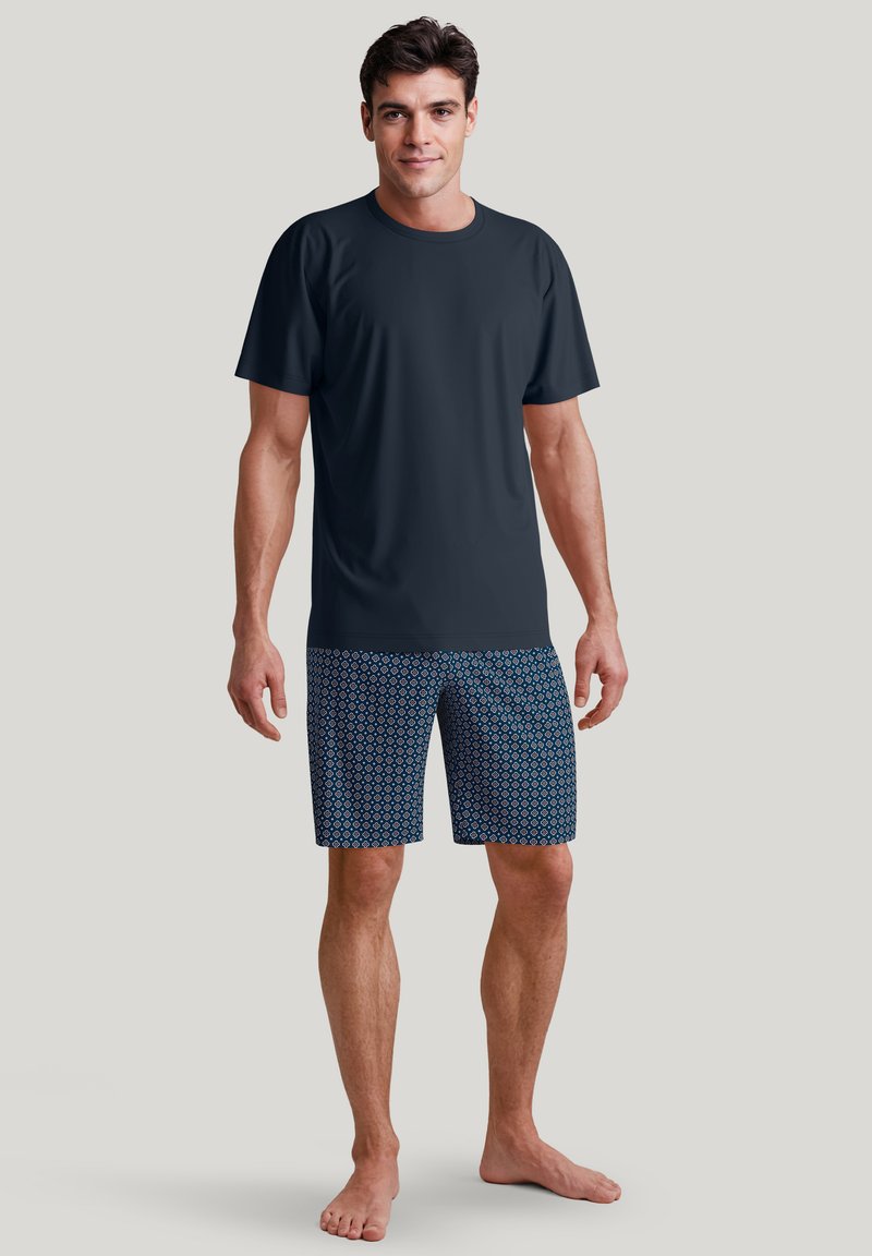 Donkere T-shirt met korte mouwen, gecombineerd met blauwe zwembroek met patronen, featuring geometrische ontwerpen. Model staat blootsvoets tegen een neutrale achtergrond.