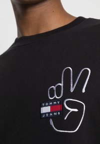 Primo piano di una maglietta nera con un simbolo della pace ricamato di colore bianco e una piccola toppa rettangolare con il logo di Tommy Jeans.