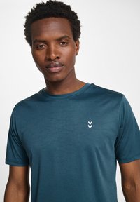 T-shirt teal à manches courtes avec un col rond et un logo flèche blanc discret sur la poitrine, fabriqué en tissu lisse et respirant.