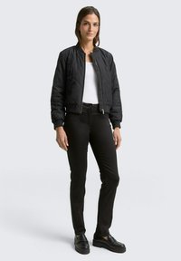 Zwarte geplooide bomberjack met een aangesloten ontwerp, ritsluiting aan de voorkant en elastische manchetten, gecombineerd met zwarte slim-fit broek en zwarte instappers.