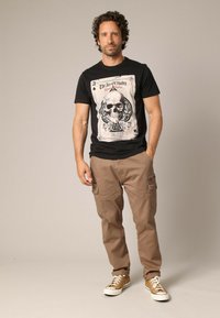 T-shirt noire avec un graphique de crâne "As de Pique", assortie à un pantalon cargo beige avec des poches latérales et des baskets marron clair.