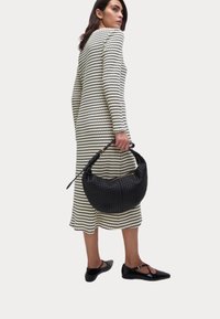 Sac à main tissé noir en forme incurvée, tenu par une personne portant une robe rayée crème et noire, assortie à des ballerines noires.