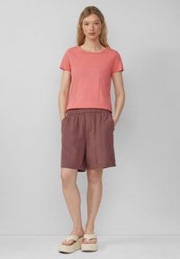 Koralrotes Baumwoll-T-Shirt mit kurzen Ärmeln, kombiniert mit braunen Leinen-Shorts mit elastischem Taillenbund und beigen Plateausandalen.
